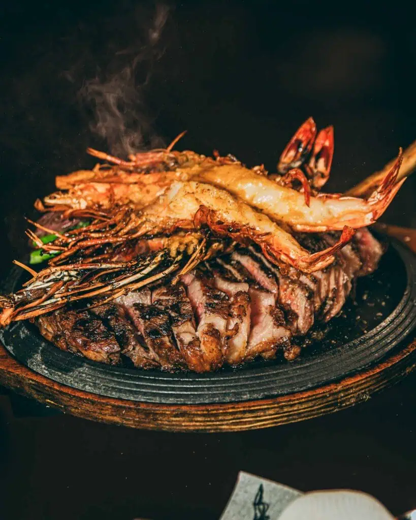 Un grand steak grillé avec des crevettes et du homard sur une plaque en fonte chaude, parfait pour une expérience de tapas espagnoles à Las Mañas.