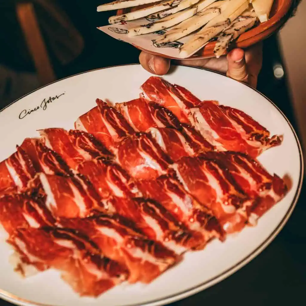 Jambon de porc avec lard et charcuterie sur une assiette blanche, avec une main tenant une assiette de tapas, adaptée aux plats espagnols, à Las Mañas.