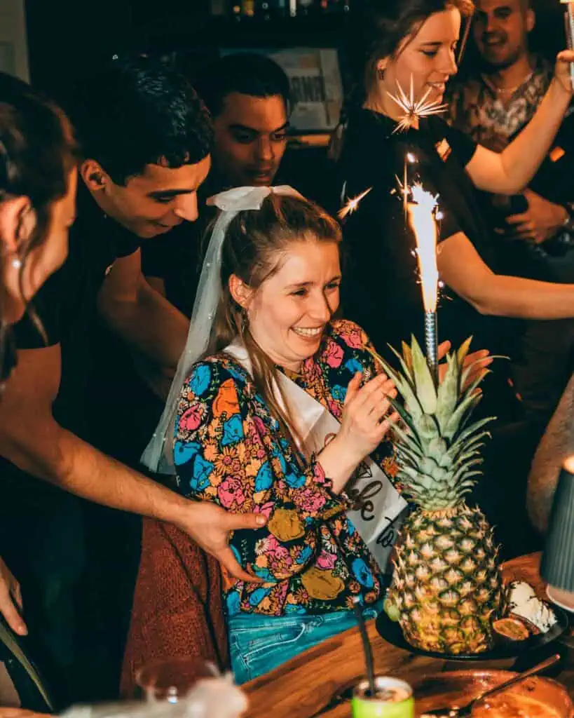 Un groupe de personnes fait la fête avec une femme qui reçoit un gâteau d'anniversaire avec un ananas et une bougie, tout en riant et en s'amusant dans une atmosphère chaleureuse.