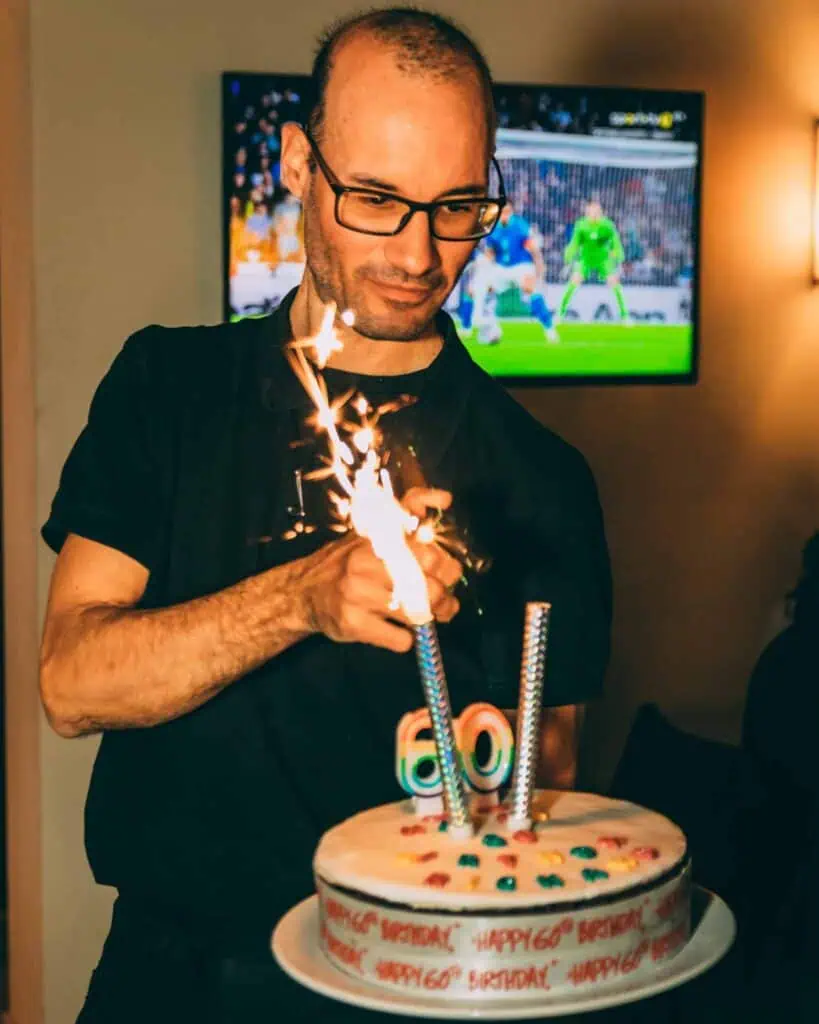 Feestelijk taart met kaarsen en sparklers voor 60e verjaardag, man die op de achtergrond sportwedstrijd op tv kijkt, gezellig verjaardagsfeest, viering verjaardag, verjaardagsdecoraties, jubileum, feestvreugde.