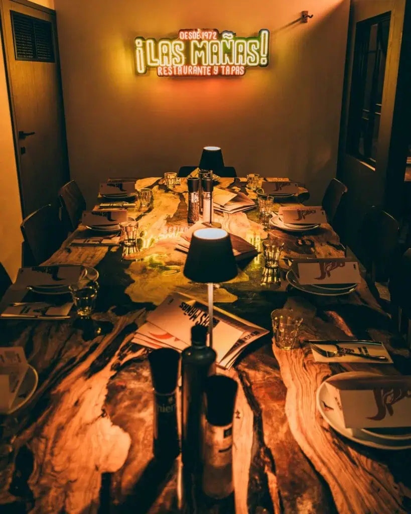 Restaurant confortable avec une grande table en bois, des chaises couvertes, des bougies de table et un néon atmosphérique appelé Las Mañas, idéal pour les tapas et la cuisine espagnole.