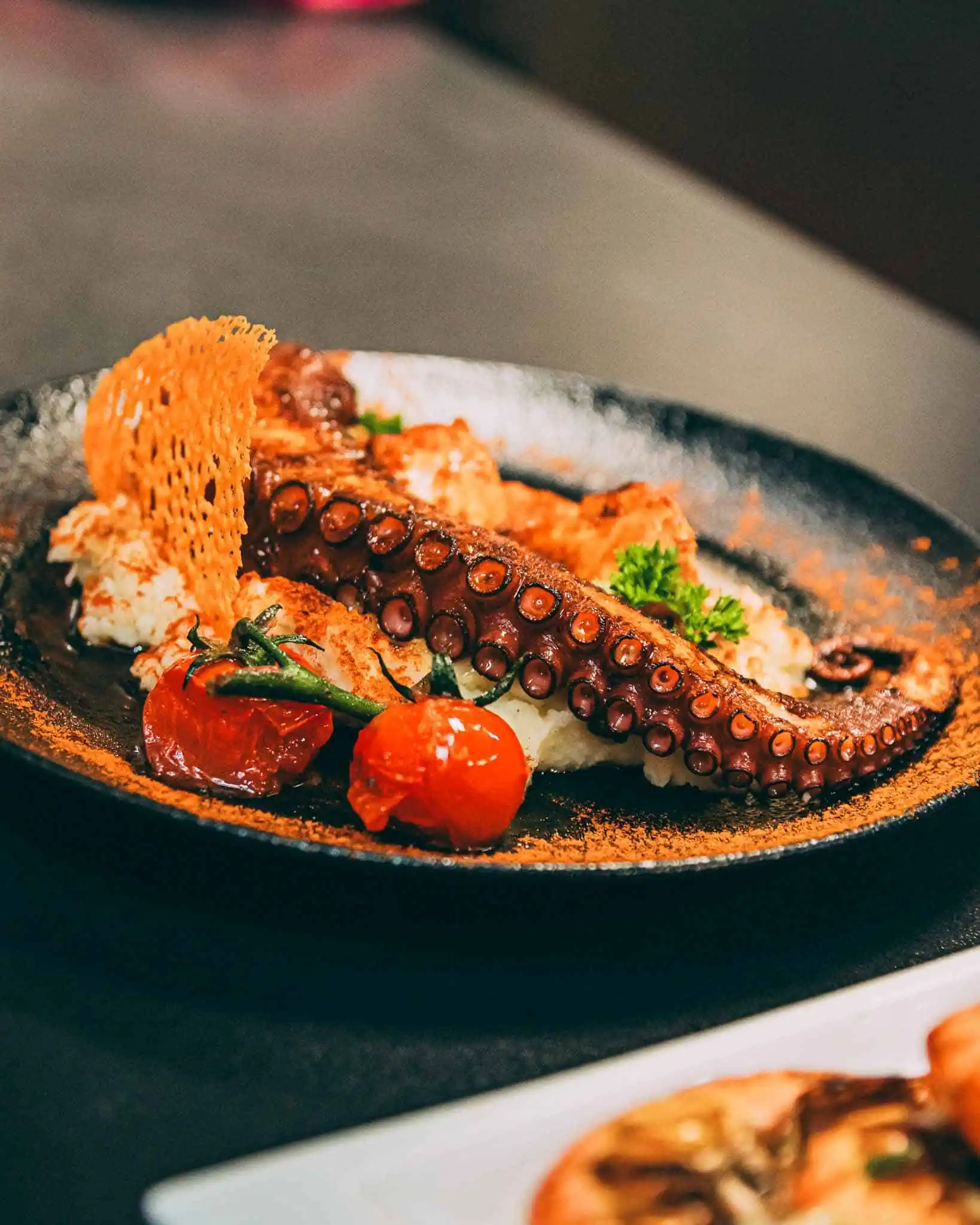 Des tapas espagnoles chaleureuses avec du poulpe grillé, des tomates cerises et des toasts croustillants sur une assiette noire, parfaites pour une expérience culinaire authentique à Las Mañas.