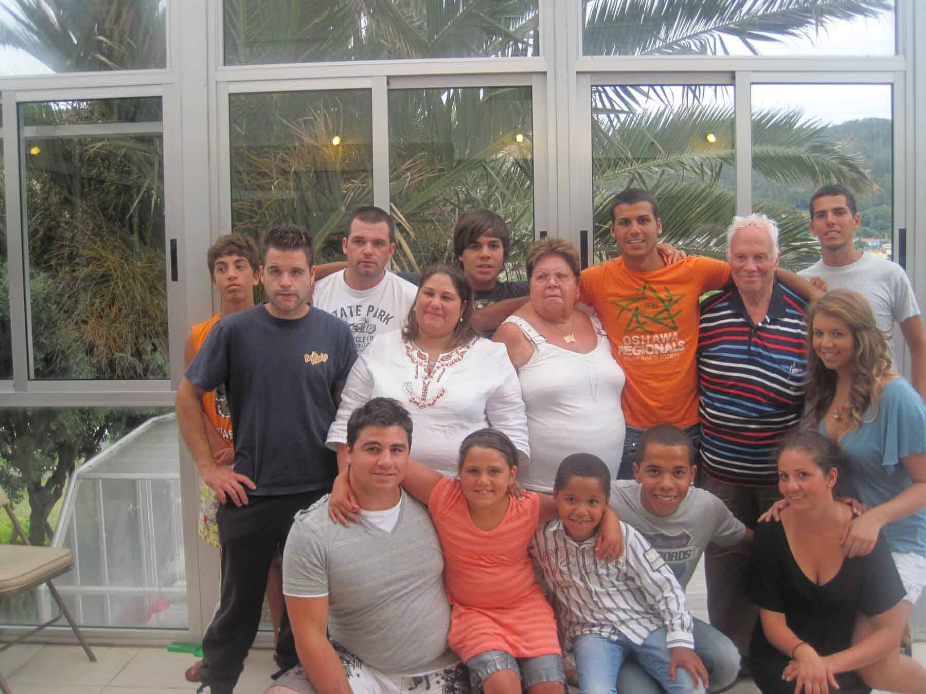 Acogedora reunión familiar o de amigos en un invernadero con grandes ventanales y plantas verdes de fondo, la gente posa junta para una foto de grupo, ambiente cálido y acogedor, luz natural y ropa colorida acentúan el sentimiento festivo.