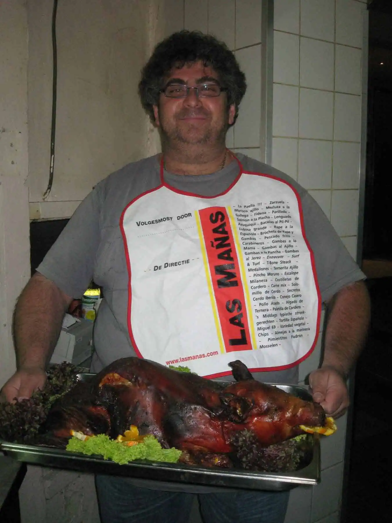Gezellige man met een glimlach houdt een groot bord met gegrild vlees en groenten, gekleed in een Las Mañas-schort met Spaans menu.