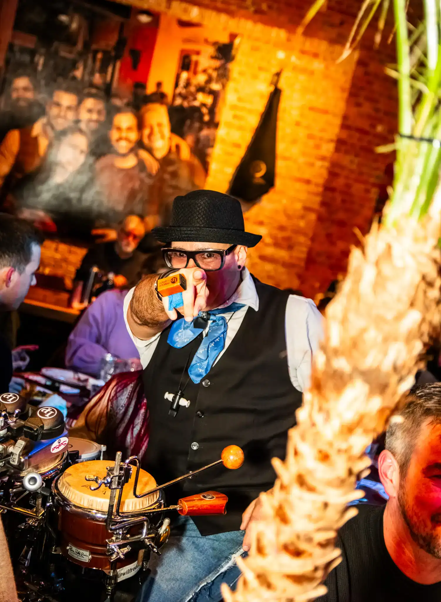 La imagen muestra a un miembro de Las Mañas Kings con un traje de fiesta con sombrero y gafas, haciendo una pose juguetona con una pistola de juguete, mientras capta el ambiente de un evento animado y sociable.